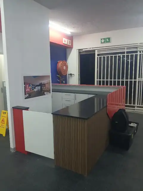 Modern L-shaped Rocket Fitness reception counter with slatted oak fluted paneling, black quartz countertops, and crimson red accent trim — Wito Projects, Johannesburg